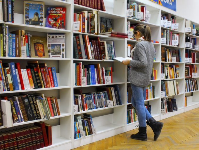 Henkilö, jolla on harmaa paita päällä ja tummat hiukset ponihännällä, tutkii kirjasto avattua kirjaa valkoisen kirjahyllyn edessä. Hyllyissä on paljon erivärisiä kirjoja.
