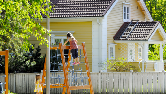 Lapsi kiipeilee kiipeilytelineessä aurinkoisena päivänä. Leikkipaikan takana on keltainen talo ja valkoinen puuaita.