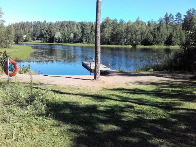 Lautaron uimaranta. Rannalla kasvaa iso puu. Rannalla telineessä pelastusrengas. Vedessä uimalaituri.