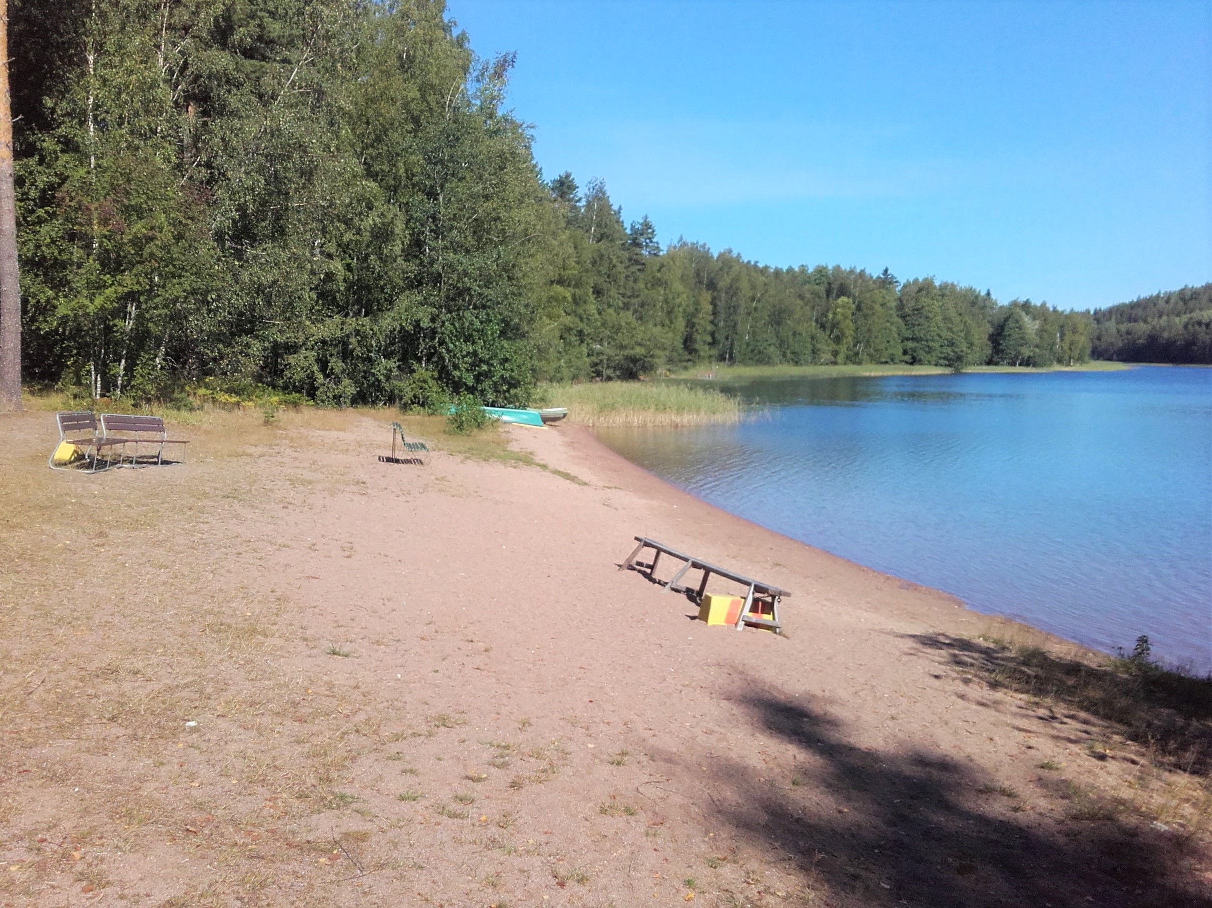 Aurinkoinen hiekkaranta. Rannalla penkkejä istumista varten. Rannan ja lammen ympärillä kasvaa metsää.