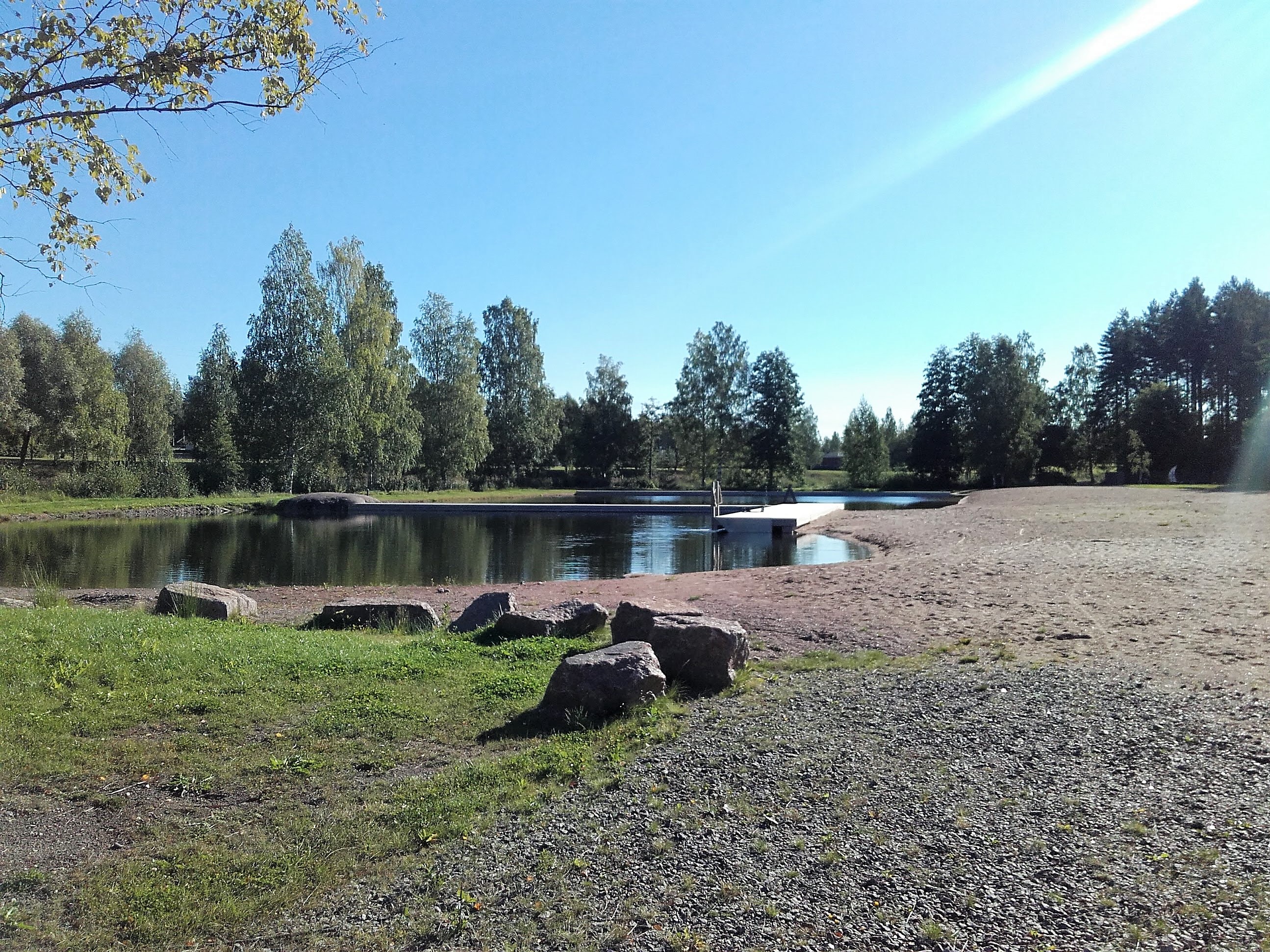 Koivusaaren maauimala. Ranta hiekkaa sekä nurmikkoa. Ympärillä kasvaa puita. Nurmikolla muutamia isoja kiviä.