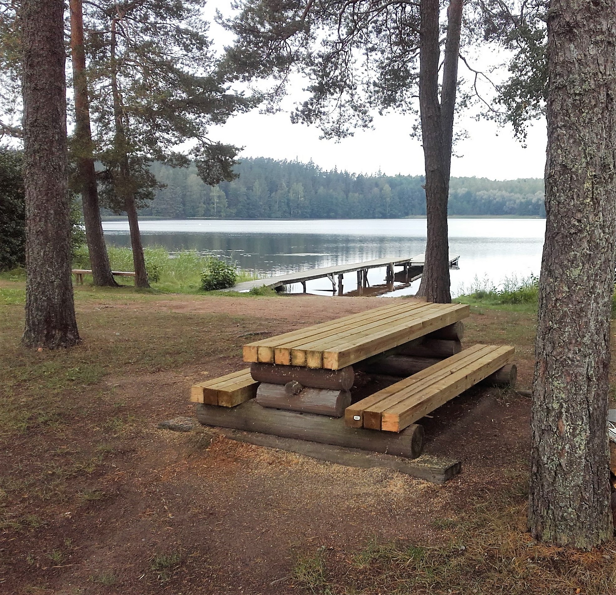 Orijärven uimaranta. Rannalla kiinteä pöytä ja penkit. Vedessä pitkä laituri. Rannalla kasvaa isoja puita.