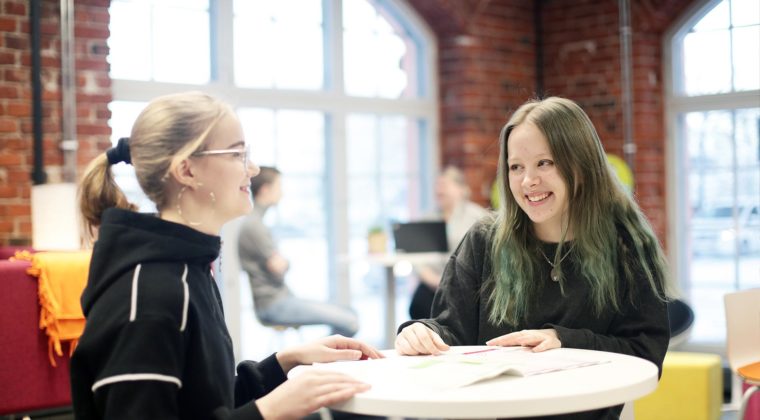 Kaksi hymyilevää nuorta naista pöydän ääressä. Tautsalla iso ikkuna ja tiiliseinää sekä muita ihmisiä.