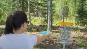 Tummahiuksinen nainen heittää frisbeetä metalliseen frisbeegolfkoriin. 