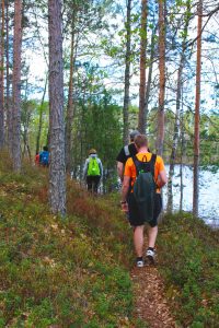 Kuvassa retkeilijöitä kävelee jonossa kuvaajan edellä polkua pitkin järven rannalla.