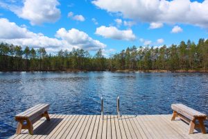 Kuvassa järvi ja etualalla laituri, jolla kaksi penkkiä ja portaat järveen. Järven toisella rannalla metsää. Taivas on kirkas ja puolipilvinen.