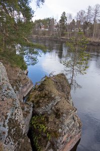 Kuvassa kallionreunaa, irtolohkare ja puita. Alhaalla näkyy sininen tyyni joen pinta.