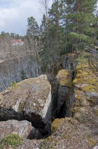 Kuvassa kallionreuna ja puita. Alhaalla näkyy sininen joki ja vastarannalla puustoa ja rakennuksia.