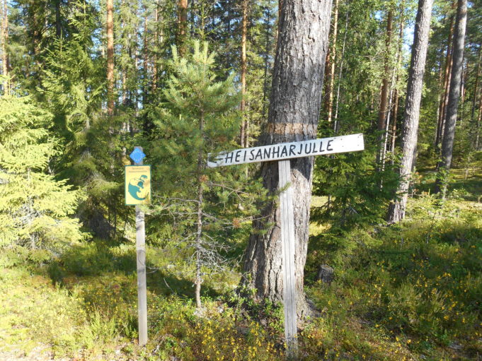 Kuvassa puinen kyltti metsässä puuta vasten. Kyltissä teksti Heisanharjulle.