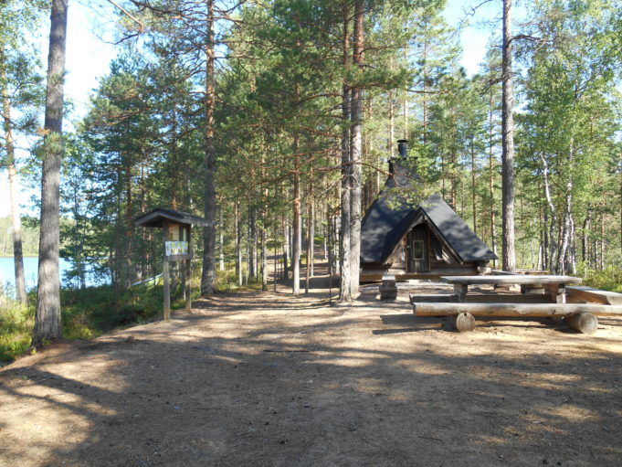 Kuvassa grillikota ja hirsistä tehty ruokailuryhmä mäntyjen keskellä. Etualalla hiekka-alue. Järvi näkyy hieman puiden välistä.