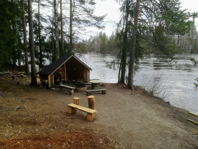 Kuvassa kota ja nuotiopaikka virtaavan joen rannalla.