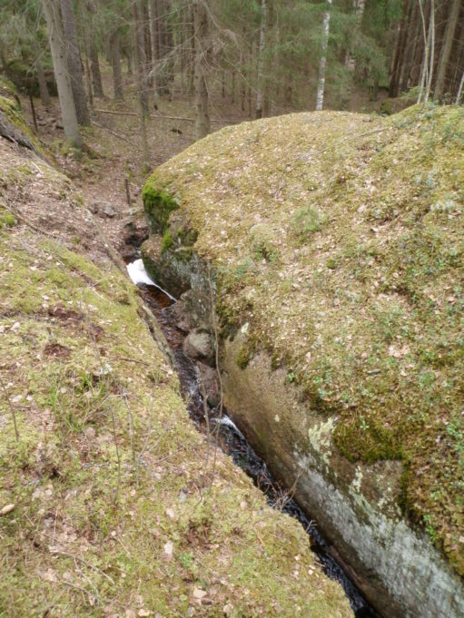 Kuvassa sammalpeitteisten kallionlohkareiden välissä kulkee puro. Taustalla kuusimetsää.