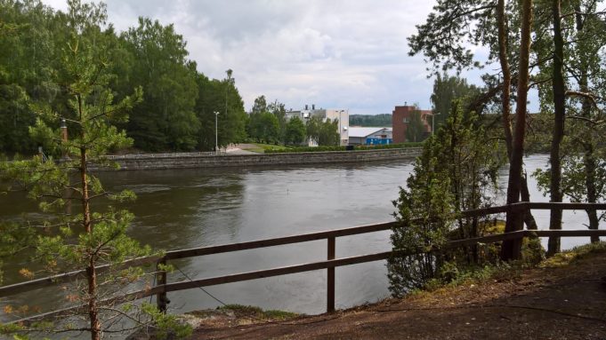 Kuvassa puinen turva-aita kallion päällä virtaavan joen rannassa. Vastarannalla metsää ja kerrostaloja.