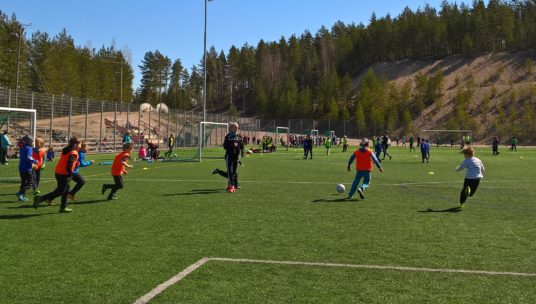 Lapset pelaamassa jalkapalloa nurmikentällä.
