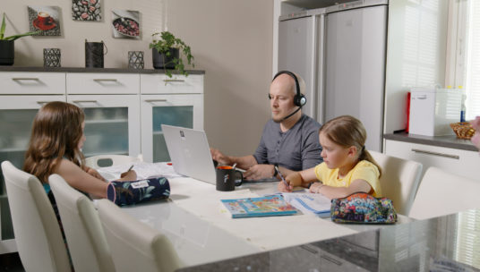 Lapset tekevät läksyjä pöydän ääressä ja isä tekee samalla töitä tietokoneella.