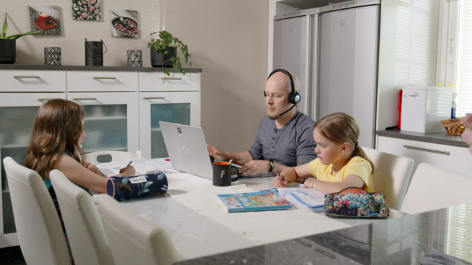 Lapset tekevät läksyjä pöydän ääressä ja isä tekee samalla töitä tietokoneella.