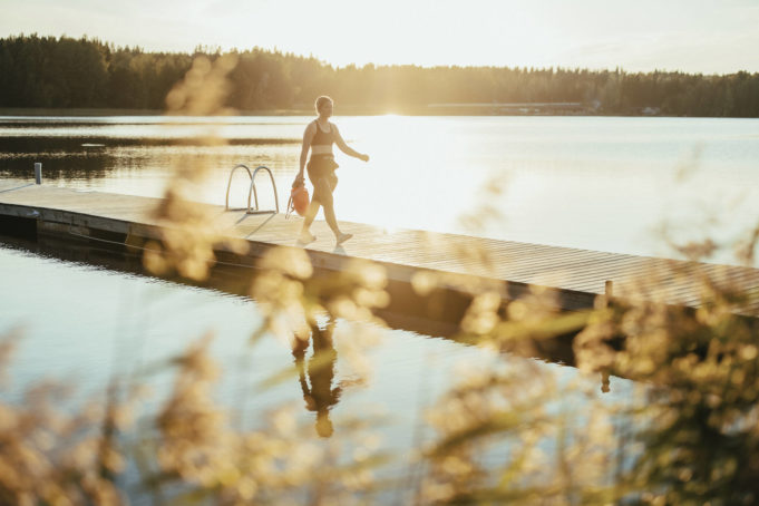 Nainen kävelee märkäpuvussa laiturilla kesäisessä järvimaisemassa.