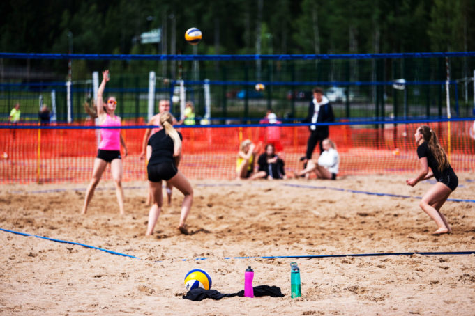 Rantalentopallon pelaajia hiekkakentällä. Pallo ilmassa. Edustalla värikkäitä juomapulloja.