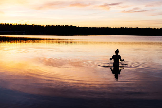 Iltakuva, nainen menossa uimaan, järvi taustalla tummaa metsää, oranssin sävyinen vesi ja taivas