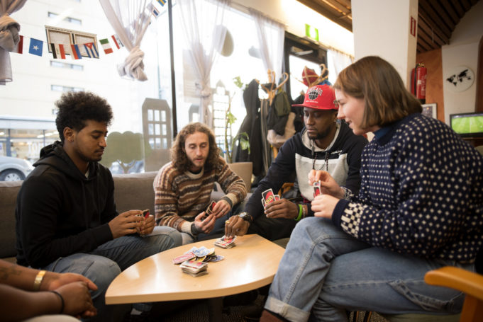 Erilaisista etnisistä taustoista tulevat henkilöt pelaavat pöydän ääressä korttia.