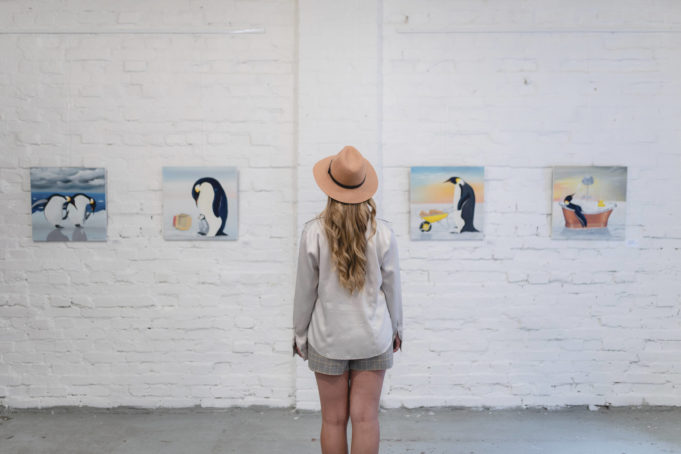 Aurinkohattuinen nainen kesäshortseissa katselee taidenäyttelyssä tauluja.