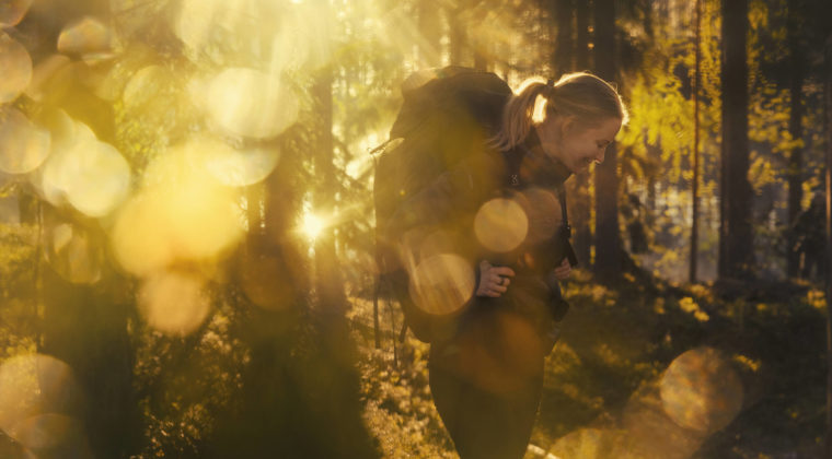 Nainen rinkkareppu selässään seisoo auringon valon kultaamassa metsässä.