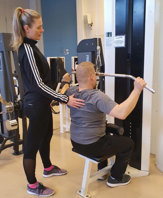 Aikuisten liikuntaneuvoja kuntosalilla ohjaamassa ylätaljavetoa.
