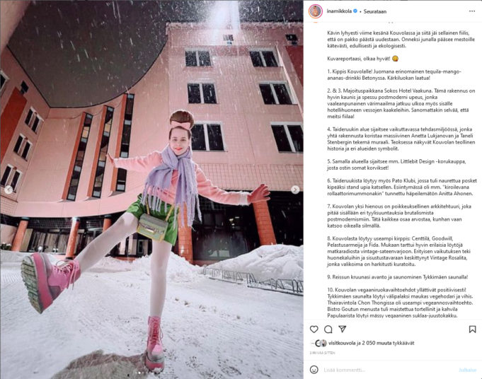 Muhkealla rusettikampauksella varustettu nuori nainen pastellisävyisissä vaatteissa seisoo yhdellä jalalla vaaleanpunaisen hotellin edessä.