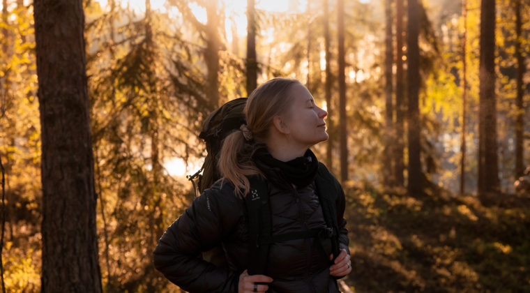 Toppatakkiin pukeutunut, poninhäntäpäinen nainen on reppu selässään syksyisessä kuusimetsässä. Laskevan auringon valo siivilöityy kuusten oksien läpi.