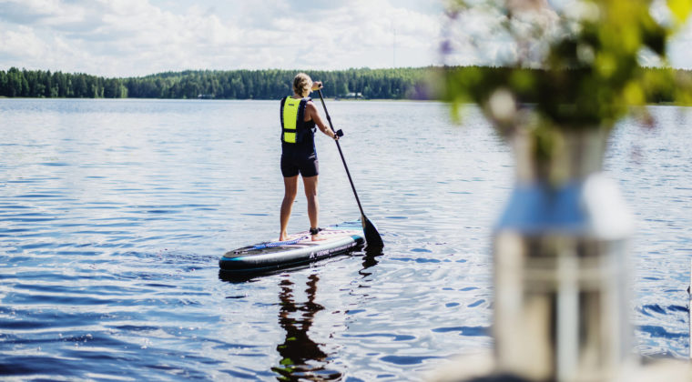 Nainen pelastusliivit päällä suplautailemassa. Kuvan edustalla maljakossa kesäkukkia.