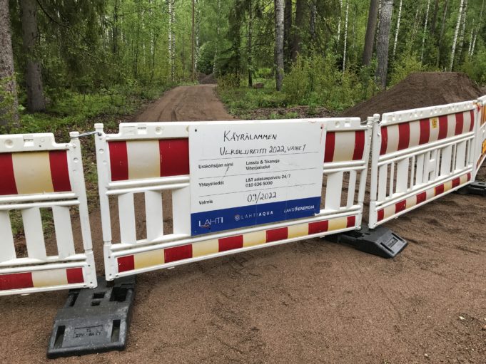 Työmaataulu ja työmaa-aita. Taustalla uudella hiekalla päällystetty ulkoilureitti ja metsää.