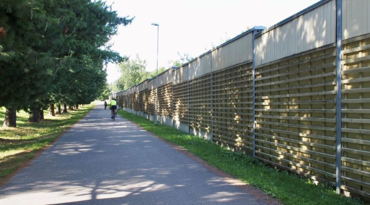 Beigen värinen, korkea meluseinä pyörätien oikealla puolella. Vasemmalla puolella puita.