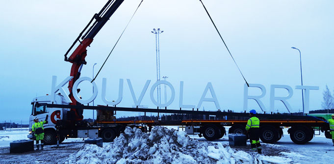 Nosturiauto siirtää suurikokoista Kouvola RRT -kylttiä paikoilleen, etualalla lumikasa.