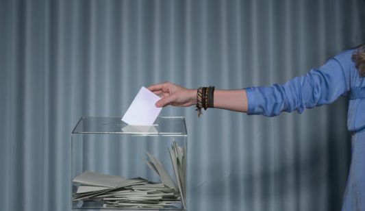 Äänestystilanne, jossa naisen käsi näkyy pudottamassa äänestyslippua läpinäkyvään vaaliuurnaan.