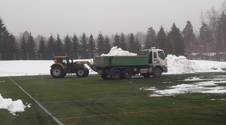 Traktori poistaa lunta tekonurmikentältä. Edustalla vihreää tekonurmea, taustalla lumikasoja ja kaksi työkonetta.