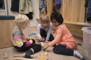 Kolme pientä tyttöä leikkii päiväkodin hyllykön edessä lelujunaradalla.