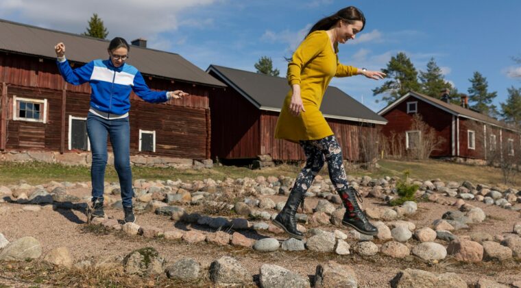 Kaksi naista kävelee kivireunuksen päällä. Taustalla vanhoja punamultarakennuksia.