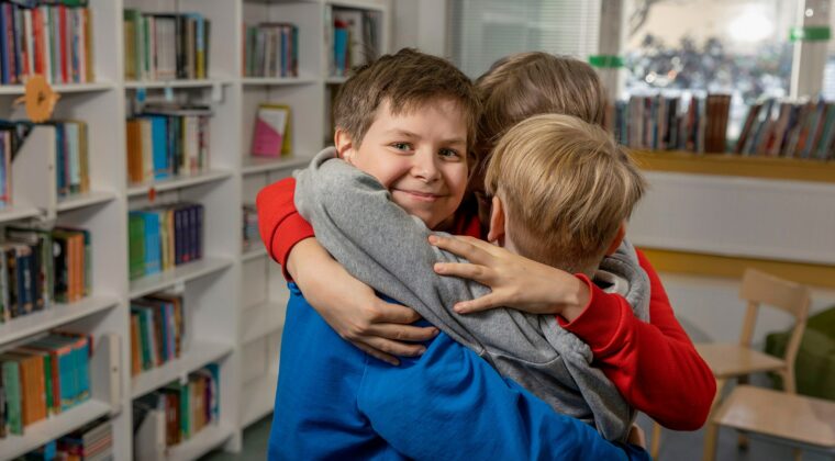 Kolme lasta halaa toisiaan. Yksi katsoo kameraa ja hymyilee. Taustalla kirjahyllyjä.