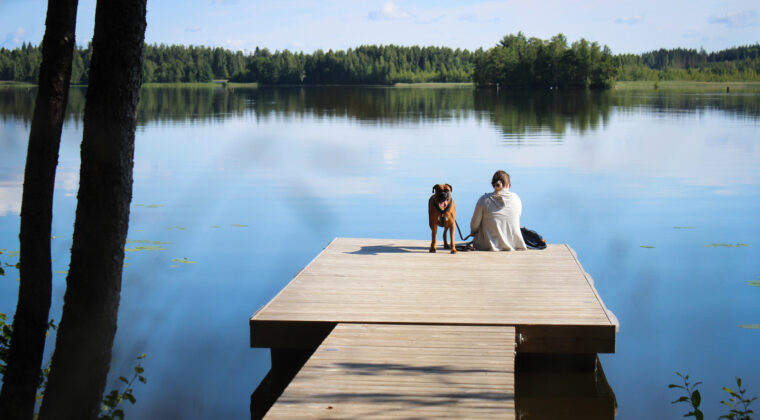 Nainen istuu selin laiturilla koira vieressään ja katselee järvelle päin. Koira katsoo kohti.