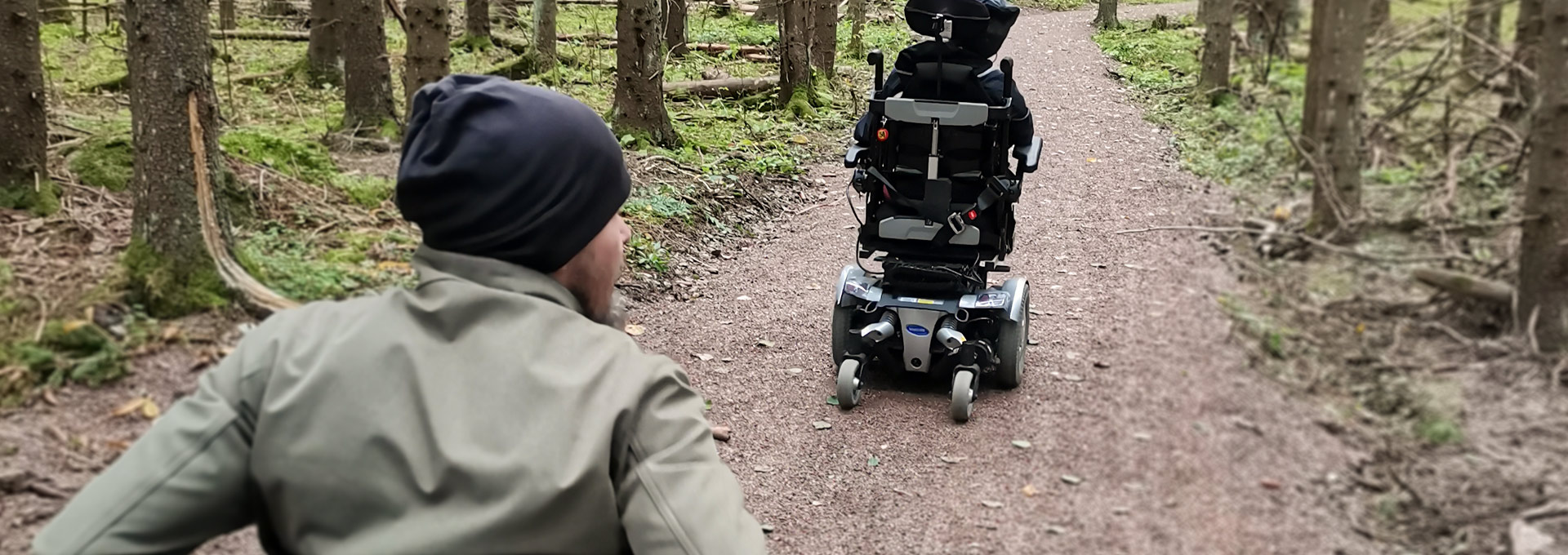 Kaksi pyörätuolin avulla liikkuvaa henkilöä kulkee hiekkatiellä metsässä.