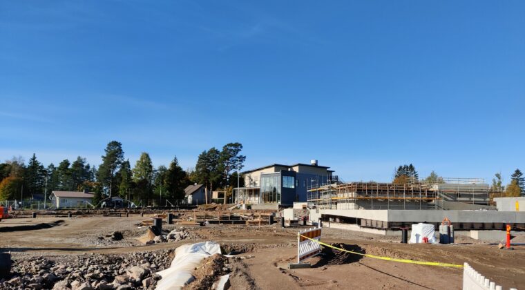 Rakennustyömaa, jossa tehdään perustuksia rakennukselle. Taustalla osa puretusta kouluta. Kirkas sininen taivas.