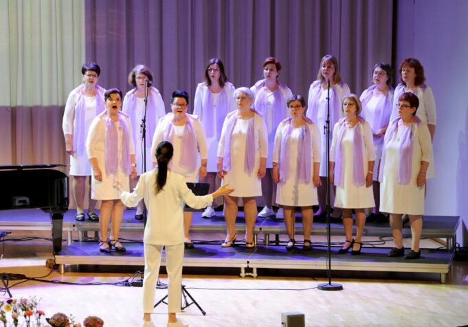 Naiskuoron naiset seisovat laulamassa kuorokorokkeilla valkoisissa mekoissaan ja kuoronjohtaja johtaa kuoroa