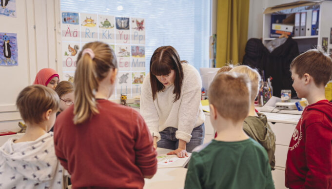 Opettaja valkoisessa paidassa näyttämässä lapsille jotain pöydällä. Oppilaat seisovat ympärillä.