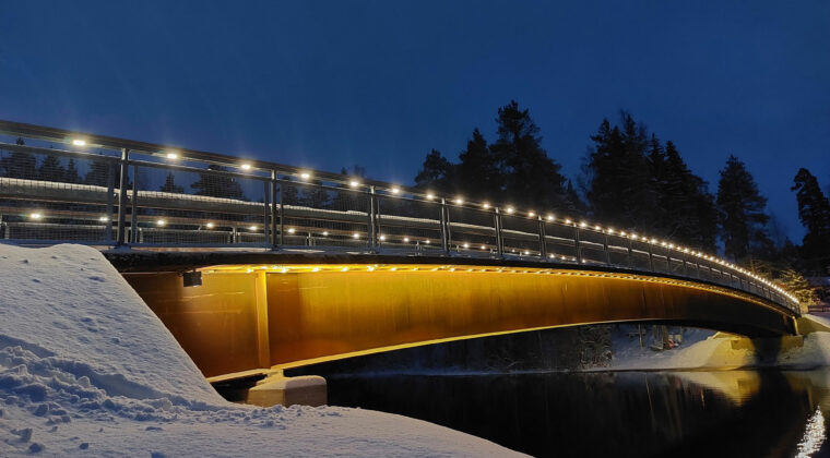 Valaistu silta joen yli lumisessa maisemassa.