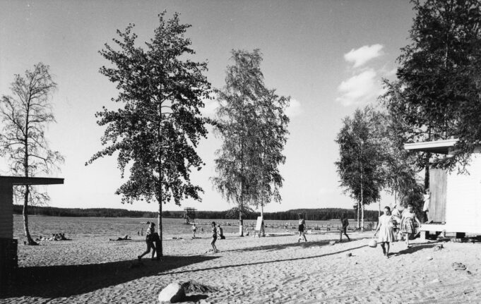 Mustavalkoinen valokuva uimarannalta, jossa makoilee ja kävelee ihmisiä. Kuvan reunoilla näkyy pukukoppirakennuksia.