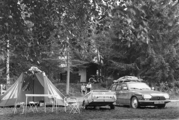 Mustavalkoisessa valokuvassa teltta, retkikalusteita, peräkärri ja auto. Taustalla pieni hirsimökki, jonka edustalla istuu kaksi henkilöä.