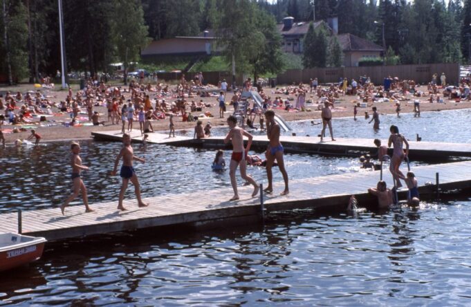 Valokuva uimarannasta, jossa runsaasti ihmisiä uima-asuissa. Edustalla iso laituri, jolla kävelee useampia lapsia. Vedessä on ihmisiä uimassa.