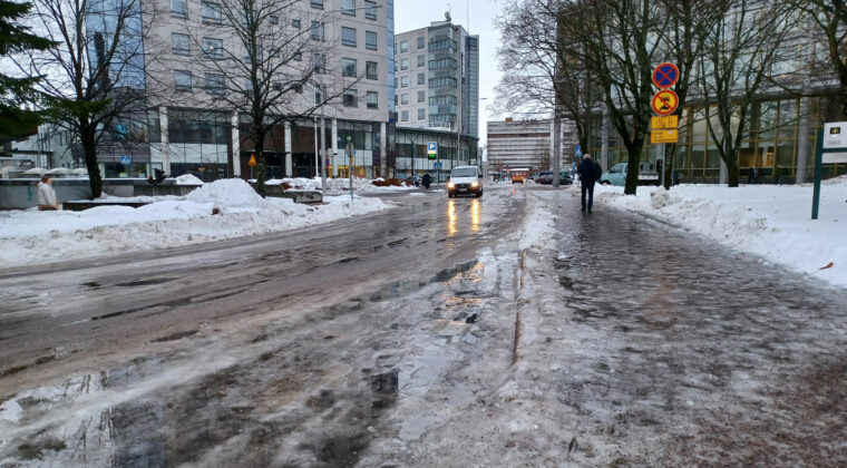 Jäinen ja märkä katu. Ympärillä kerrostaloja. Auto ja yksittäinen kävelijä kadulla.