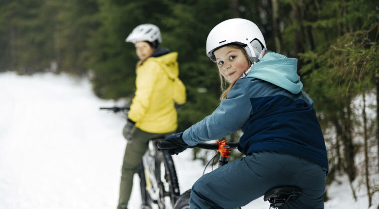 Nuori pyöräilijä valkoisessa kypärässä katsoo vasemman olkapään yli kuvaajaa. Edessä toinen pyöräilijä keltaisessa takissa. Talvi, märkä ja harmaa sää.