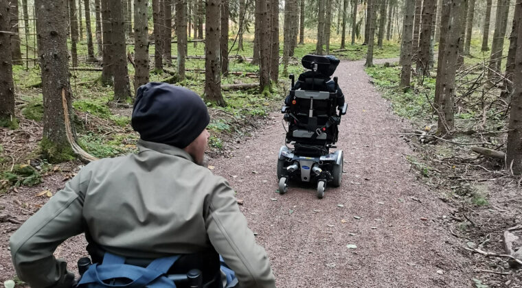 Kaksi henkilöä liikkumassa pyörätuoleilla ulkoilureitillä metsässä.